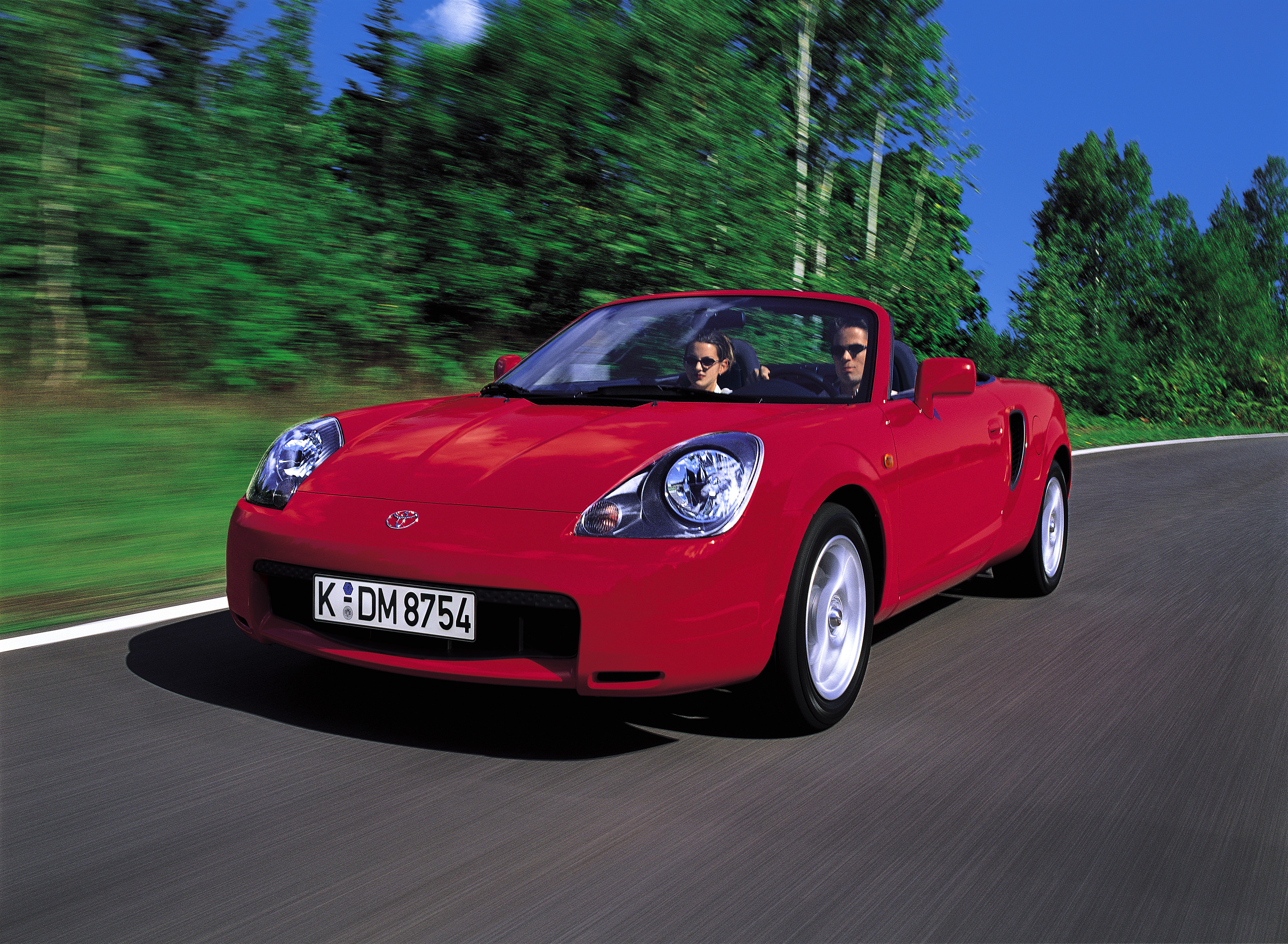 Roter Toyota MR2 W30 Roadster fährt frontal auf einer Straße mit verschwommenem grünen Hintergrund, zwei Personen an Bord.