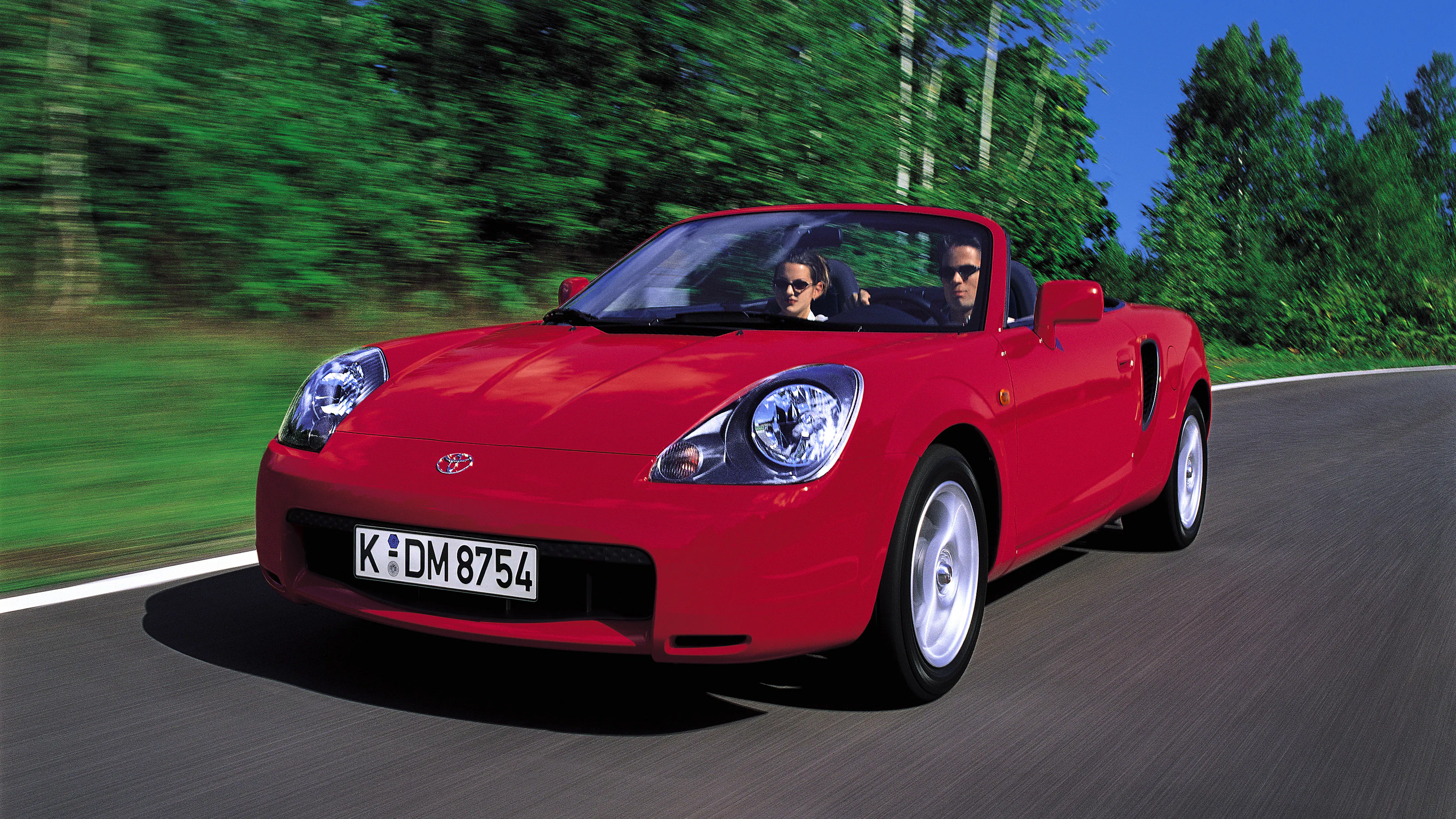 Roter Toyota MR2 W30 Roadster fährt frontal auf einer Straße mit verschwommenem grünen Hintergrund, zwei Personen an Bord.