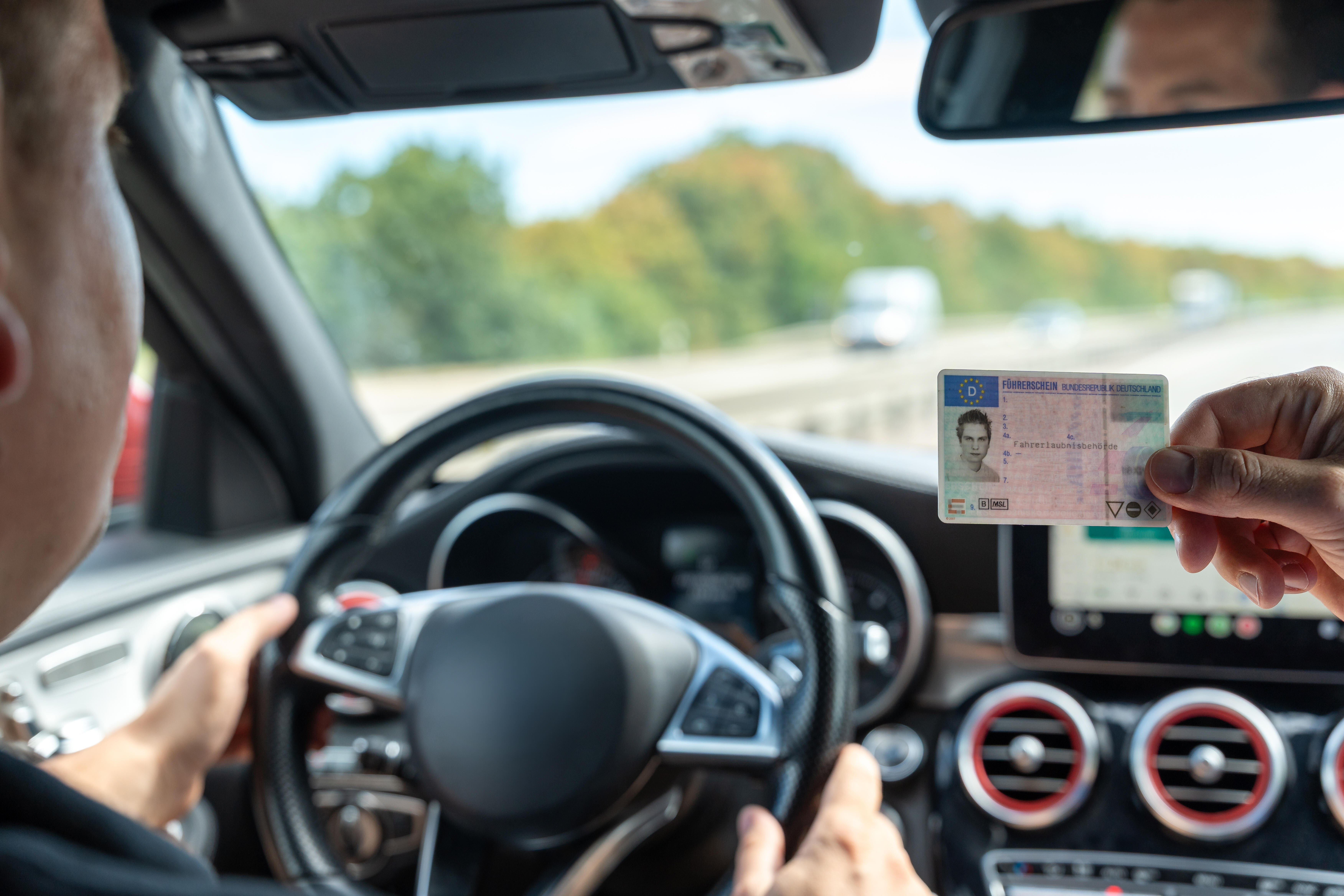 In einem Fahrschulauto sitzt ein Fahrschüler am Steuer. Von rechts hält jemand einen Führerschein ins Sichtfeld.