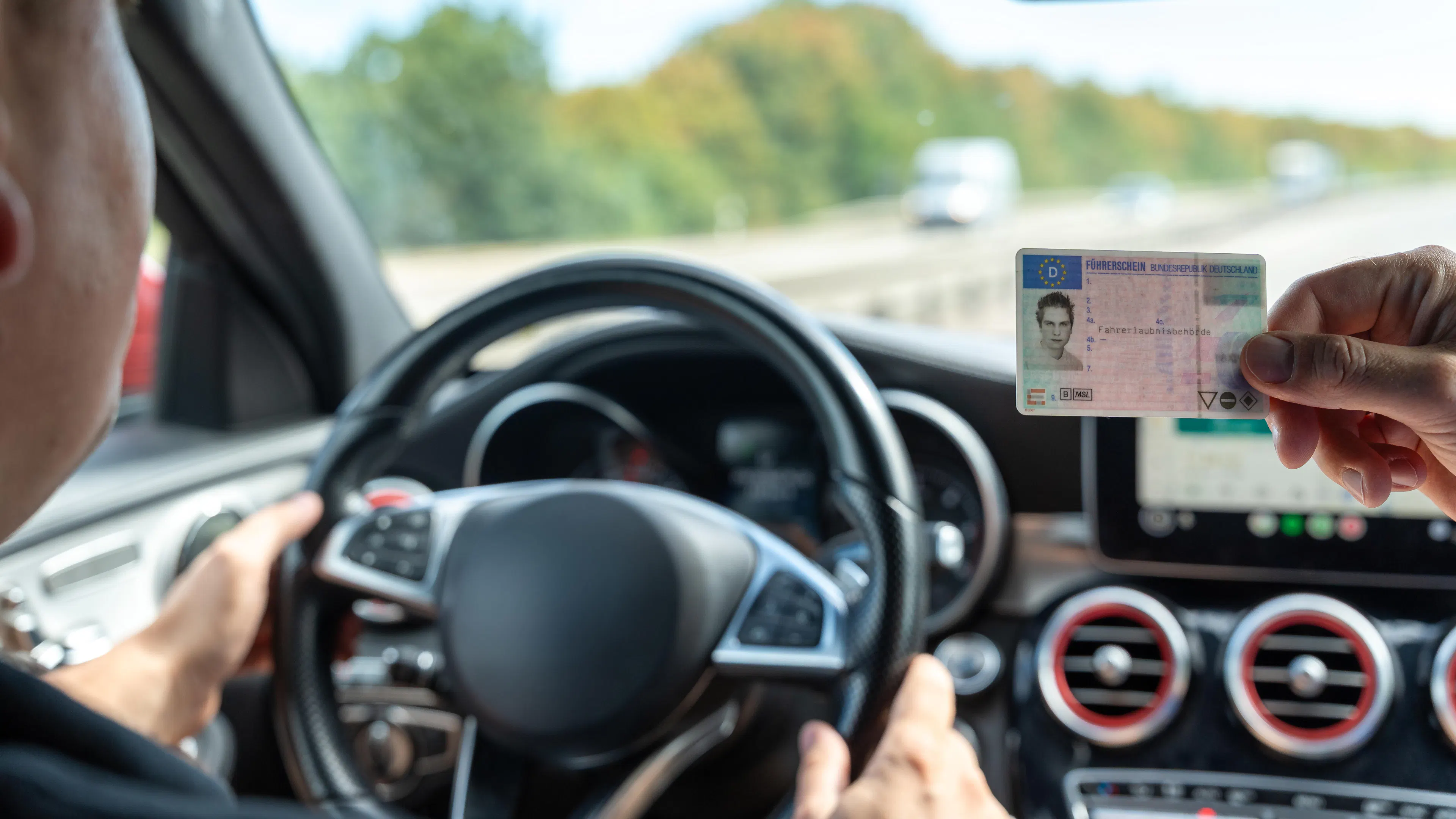 In einem Fahrschulauto sitzt ein Fahrschüler am Steuer. Von rechts hält jemand einen Führerschein ins Sichtfeld.
