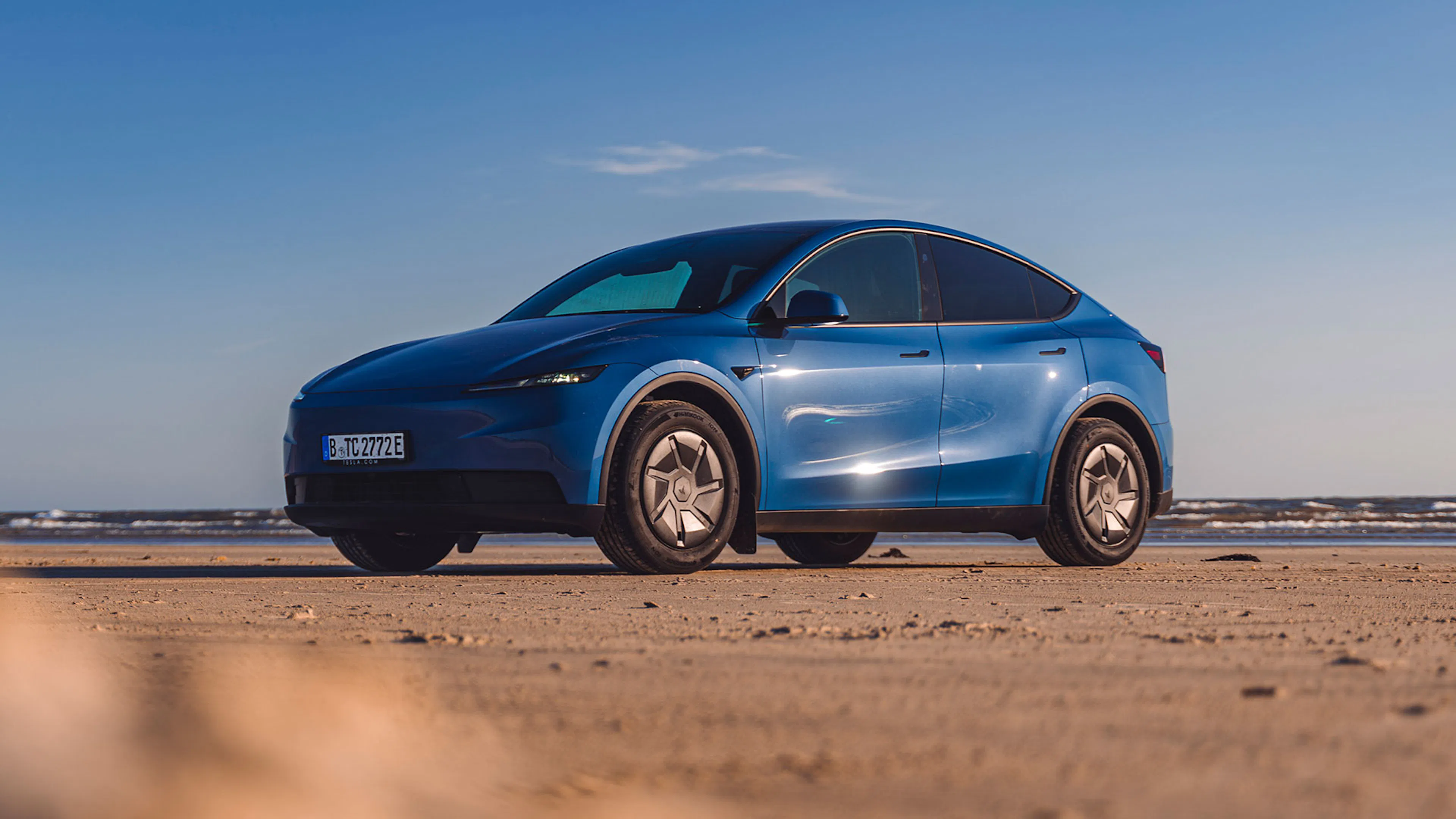 Ein blaues Tesla Model Y steht am Strand, im Hintergrund branded das Meer in kleinen Wellen an den Strand.