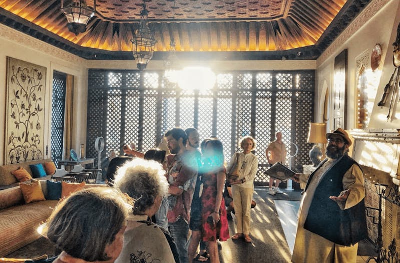 Photograph of past resident Abdul-Rehman Malik standing in front of the fireplace in the living room during sunset. Visitors are standing and looking at collection pieces. 