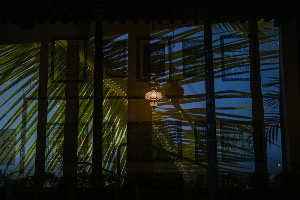 still image of Aina Paikai's video artwork for 8x8_shangrila_23. Projected image of palm fronds onto the wall of Shangri La's central courtyard at night. a single hanging lamp is turned on in the middle of the courtyard.