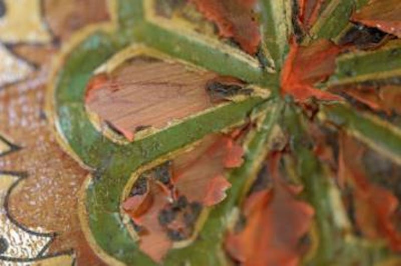 Detail of the flaking paint in a flower motif on the foyer ceiling. The red is the ground on the underside of the presentation surface. The presentation surface was gilded but is now tarnished and appears black.