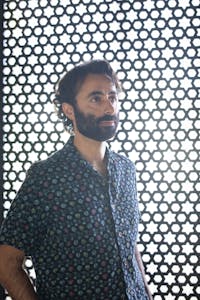 Artist, Navid Sinaki, standing up looks upward. He is standing in front of a jali wooden screen with light coming through. 