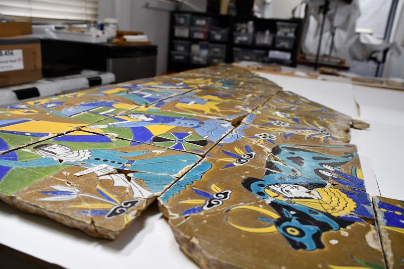 Photo of ceramic tiles in conservation lab. Two figures on tiles. Male figure crouching on the left wearing a real rob. Other parts of the scene are not easily visible. 