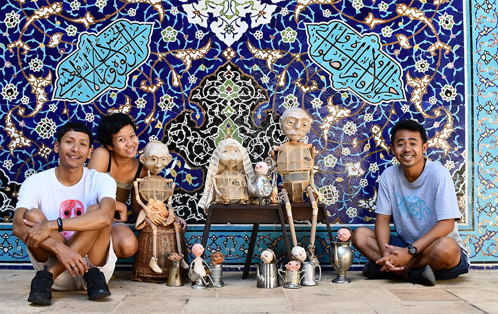 Photo of three people from Papermoon Puppet Theatre with their. Three puppets are sitting on stools, eight puppets are located on the floor. People and puppets are posing in front of a mosaic.