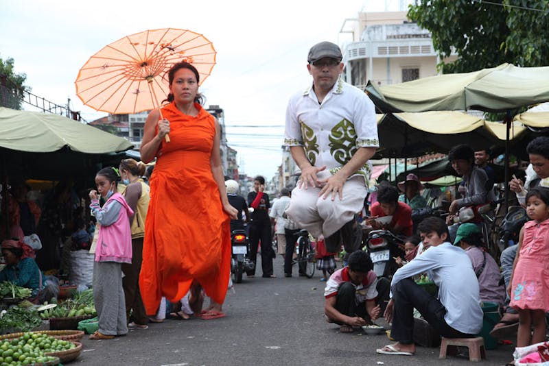 Woman wearing a bright orange dress and holding 
 an orange parasol while jumping in the air with a man wearing a hat and white shirt, also jumping in the air so that it appears that they are floating. 