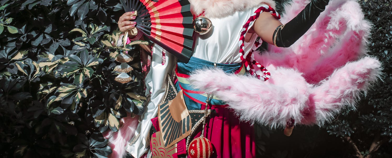 Myleah in cosplay as Karatoga Tenn from IDOLiSH7. Photo by Ubevisuals. The image is cropped from the shoulder to mid-thigh. Myleah wears a white shirt an red flowy pants with many, colorful belts. She's holding a red-and-black fan. An apple-sized jingle bell hangs from her neck. A large, pink, and fluffy multi-pronged tail brings the costume together.