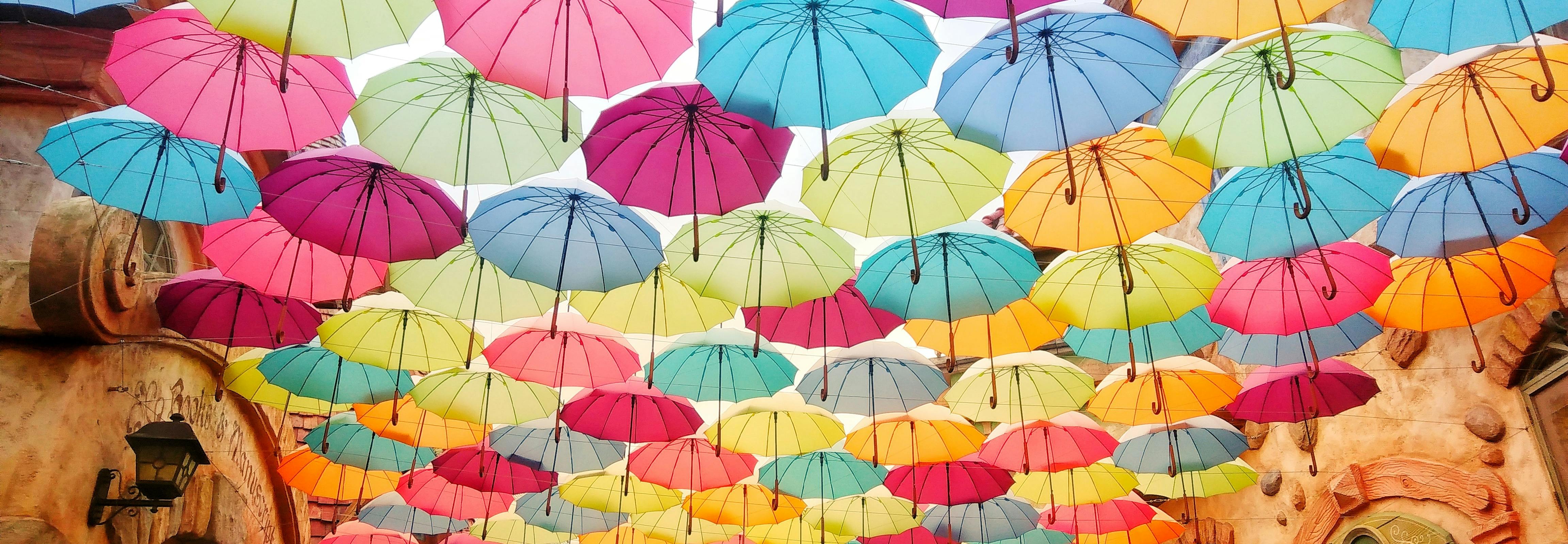 Rows of colorful umbrellas hang and form a roof above an old-timey street. Light shines from between and through the umbrellas, casting the street in a soft light. Photo by Helen Cheng on Unsplash.