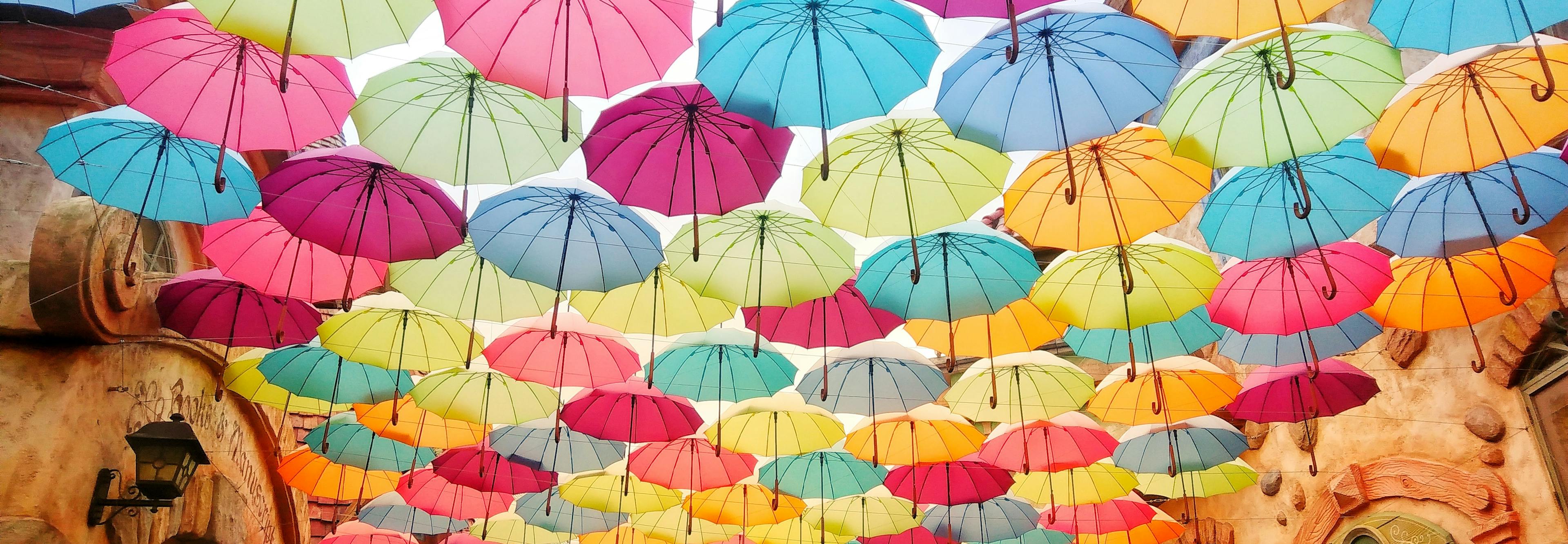 Rows of colorful umbrellas hang and form a roof above an old-timey street. Light shines from between and through the umbrellas, casting the street in a soft light. Photo by Helen Cheng on Unsplash.