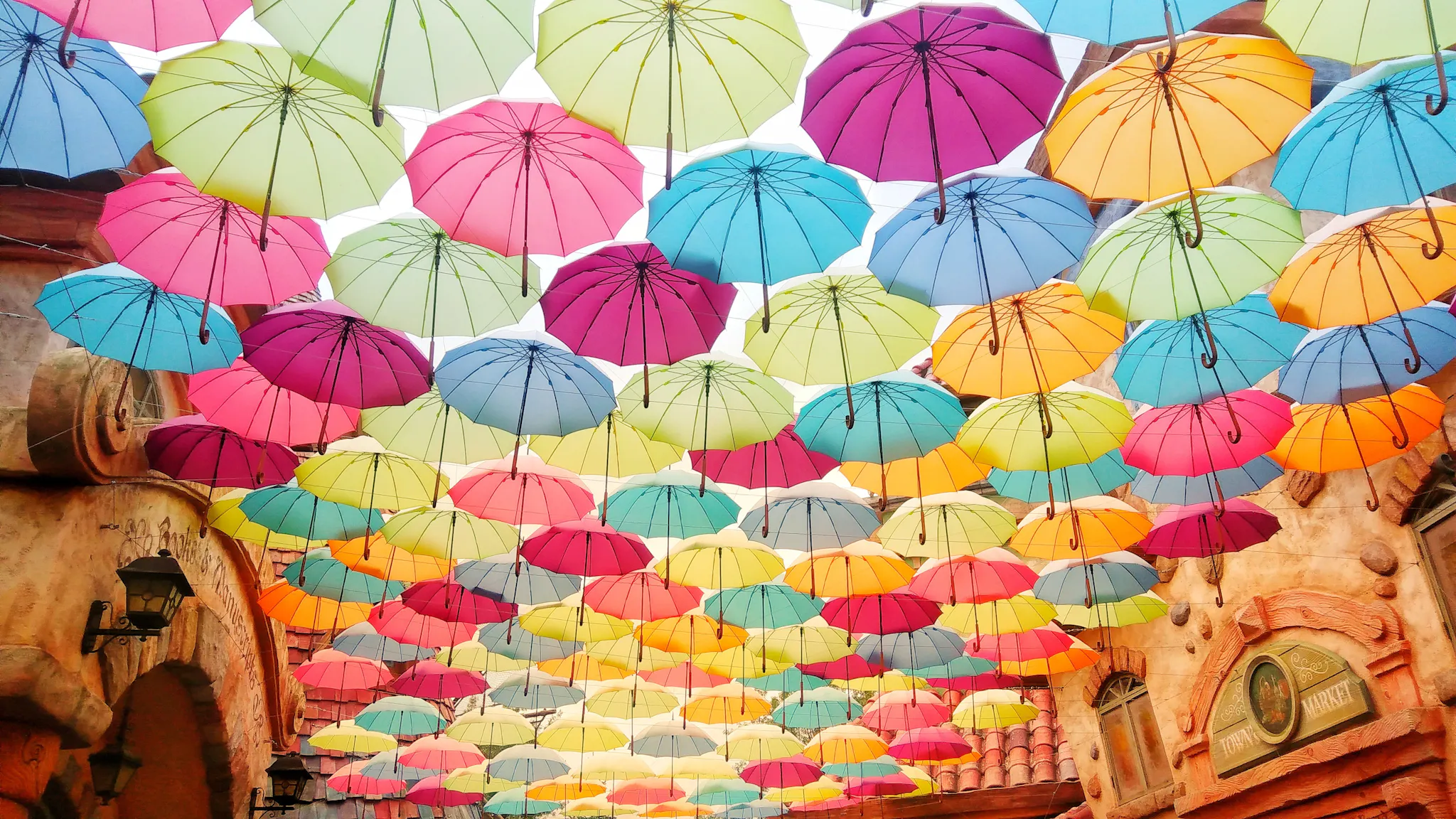 Rows of colorful umbrellas hang and form a roof above an old-timey street. Light shines from between and through the umbrellas, casting the street in a soft light. Photo by Helen Cheng on Unsplash.