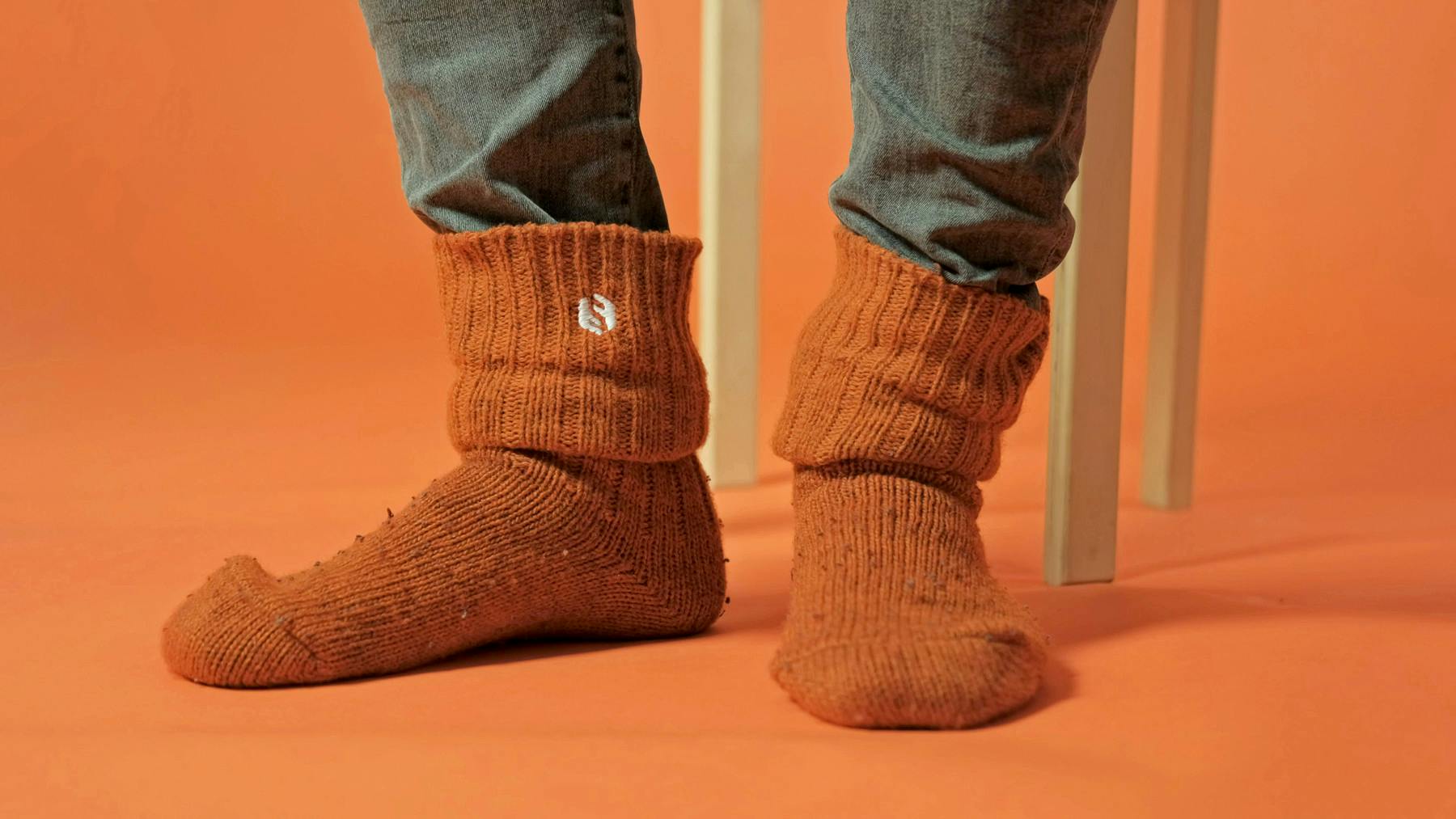 Two feet wearing Sharetribe wool socks on an orange background. Behind the feet, legs of a wooden chair.