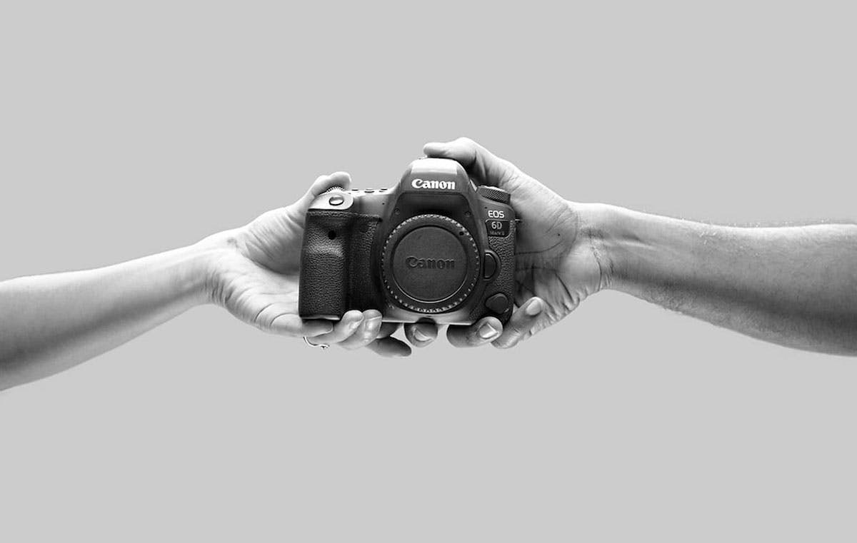A black-and-white image two hands holding a Canon camera. The hand on the left is offering the camera to the hand on the right.
