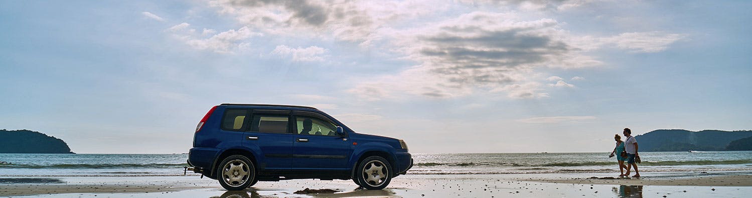 Two people walking from the right towards a blue SUV parked on a beach.