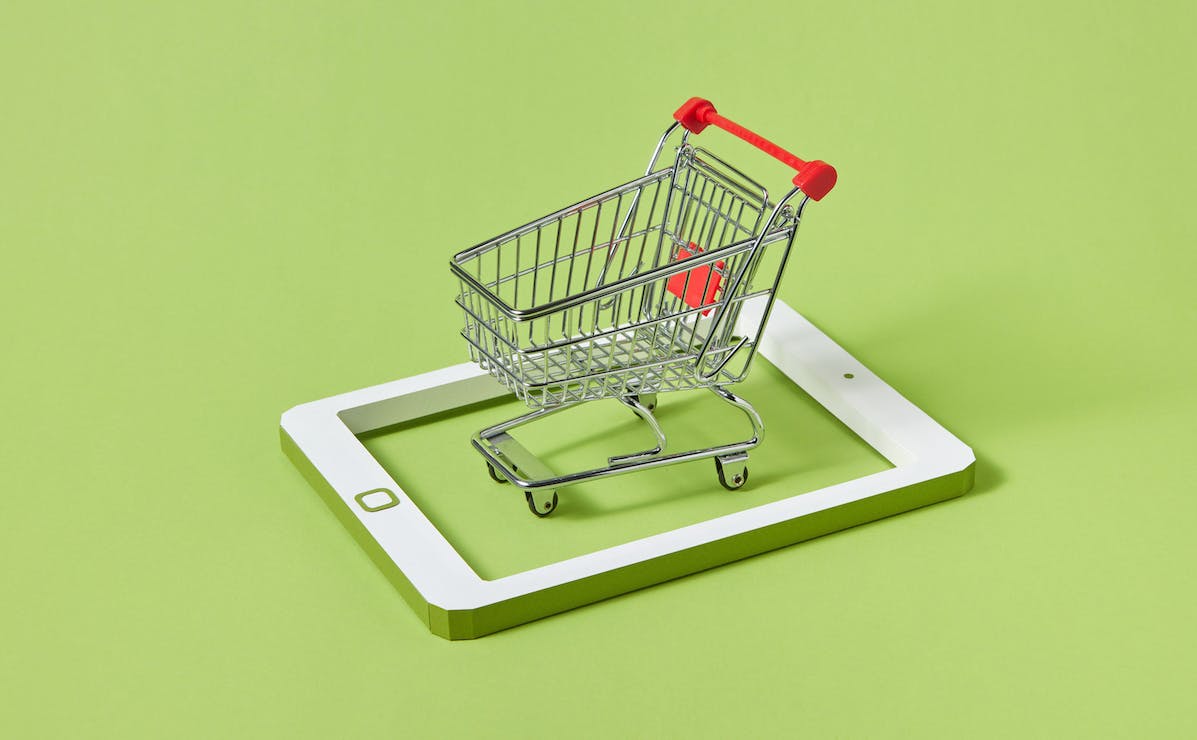 An empty miniature shopping in the middle of a tablet made out of white and green cardboard. The table computer does not have a screen. It's just the frame, lying on a smooth green surface..