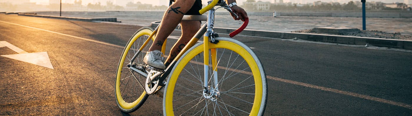 Person riding a yellow-wheeled bike on asfalt road next to a beach at sunset, image cropped to show only biker's legs.