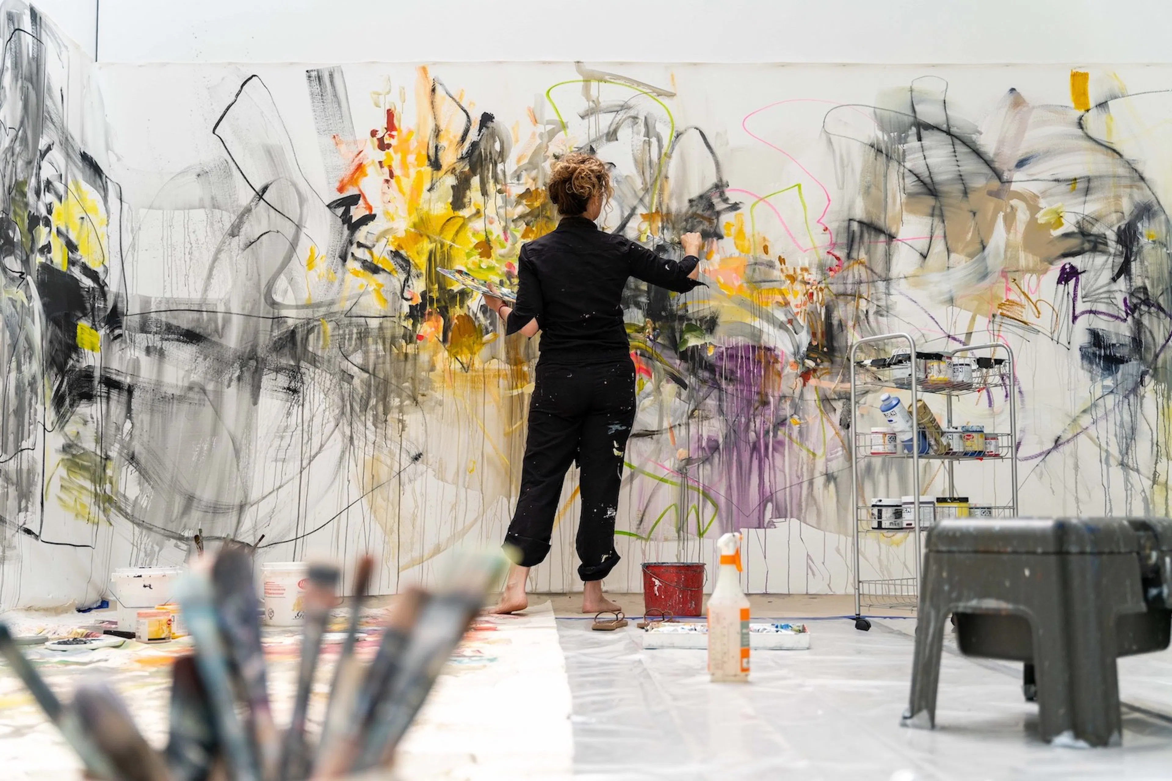 A barefoot artist, dressed in black is photographed from the back, working on an abstract painting that fills the entire wall.