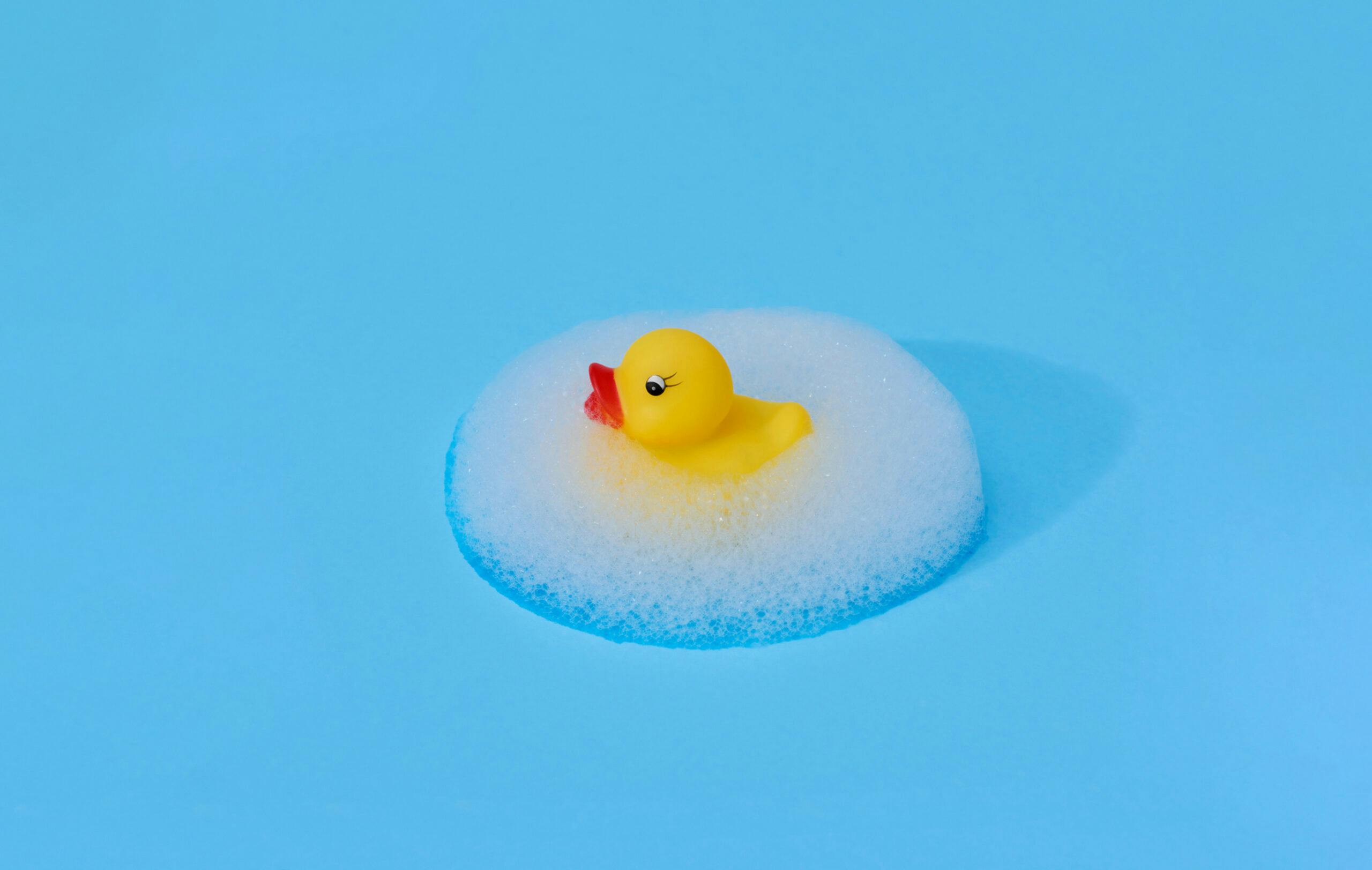 Yellow rubber duck surrounded by a symmetrical ellipsis of foam against a bright blue background.