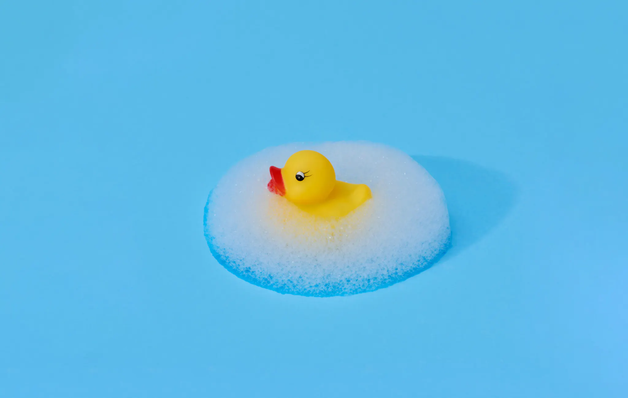 Yellow rubber duck surrounded by a symmetrical ellipsis of foam against a bright blue background.
