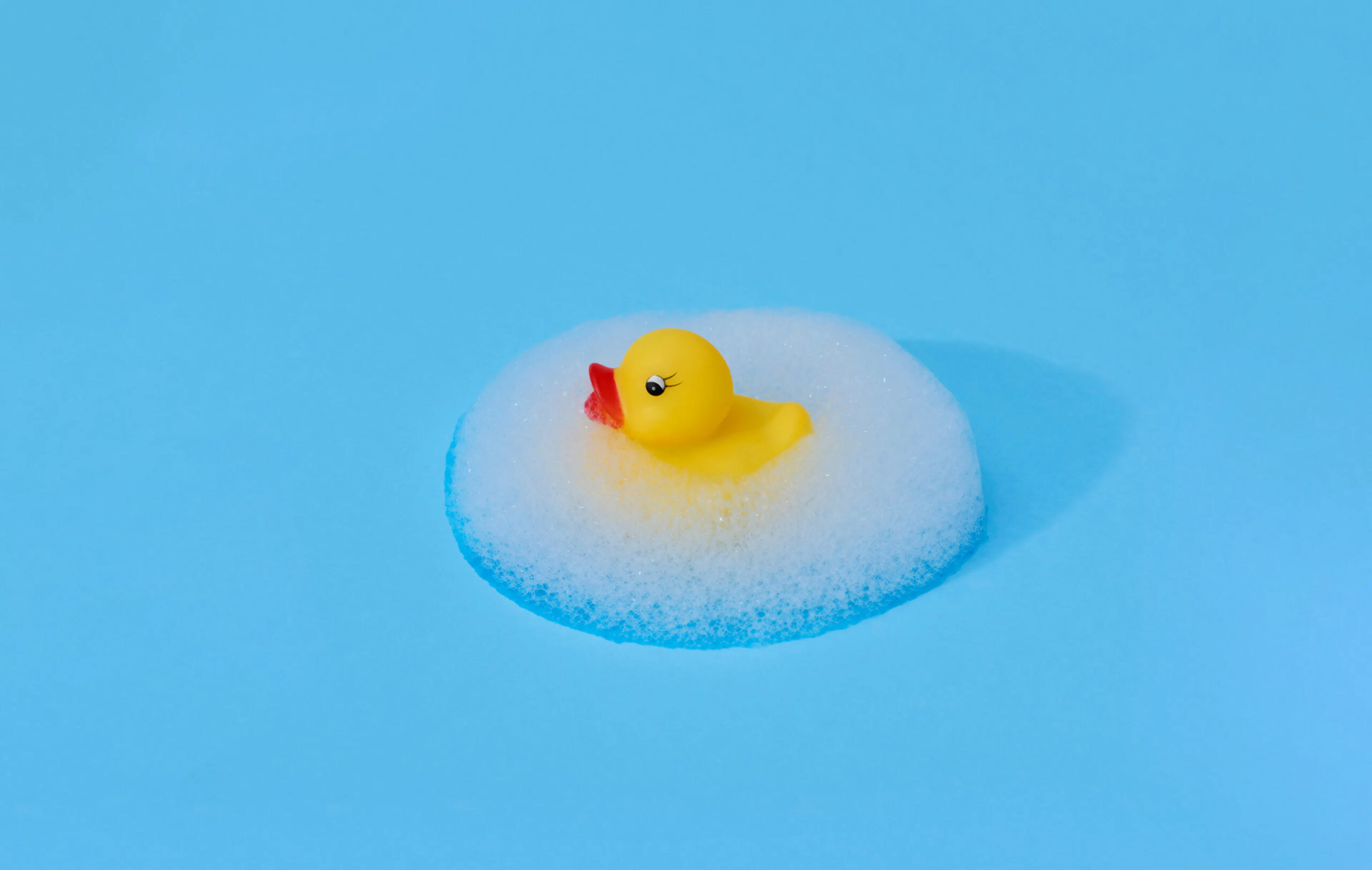 Yellow rubber duck surrounded by a symmetrical ellipsis of foam against a bright blue background.