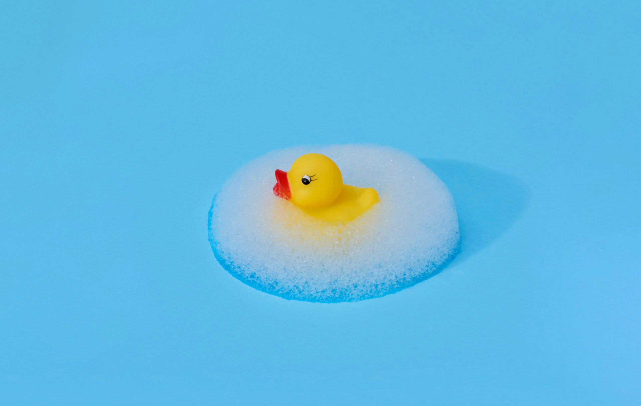 Yellow rubber duck surrounded by a symmetrical ellipsis of foam against a bright blue background.