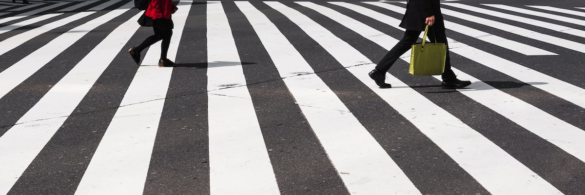 Two people walking across a wide zebra crossing. Only their legs are visible. One is wearing a red jacket and a skirt, the other has black trousers and a green bag.