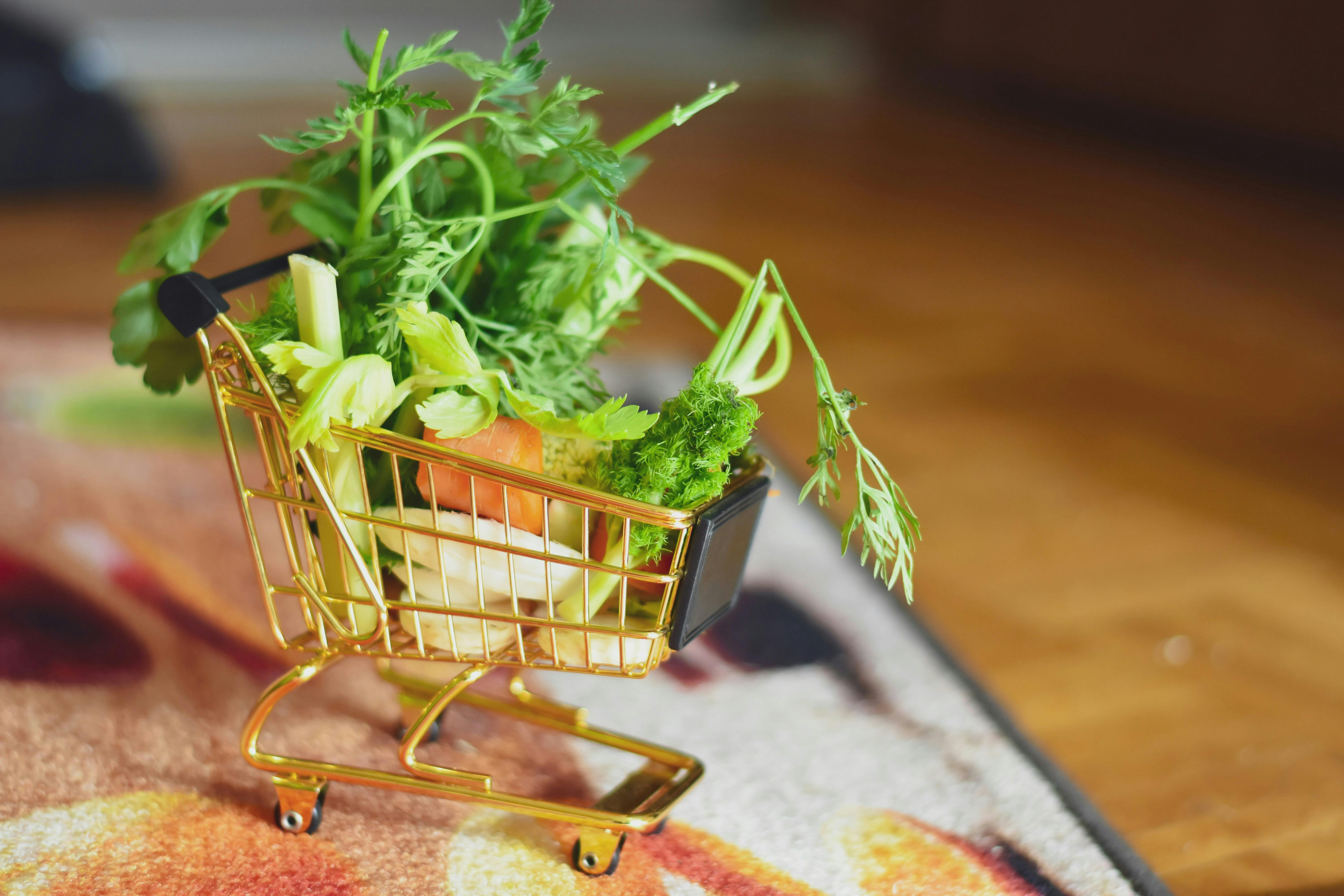 A small brass shopping cart overflowing with small vegetable parts on a soft rug, with some wooden floor visible next to the rug.