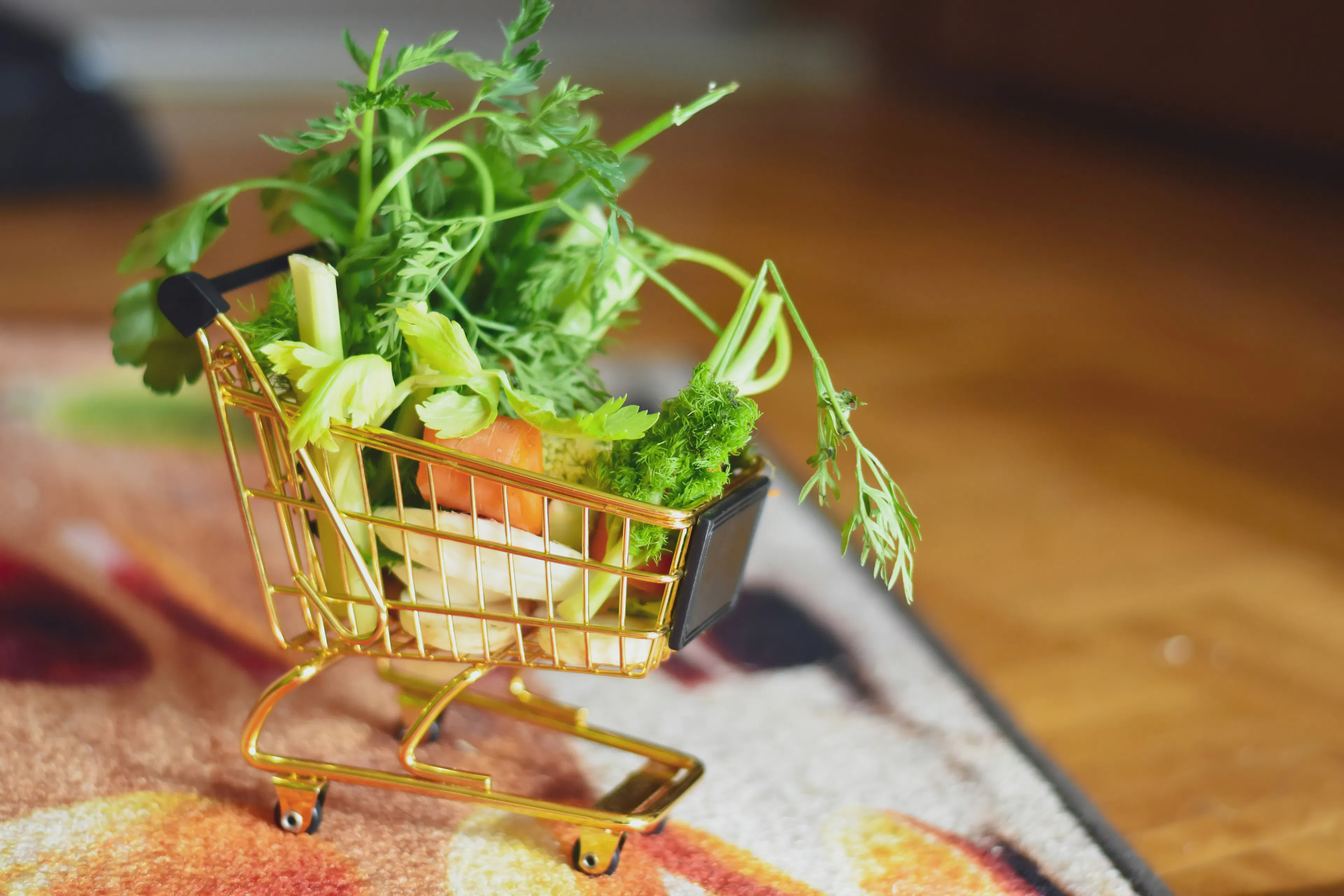 A small brass shopping cart overflowing with small vegetable parts on a soft rug, with some wooden floor visible next to the rug.