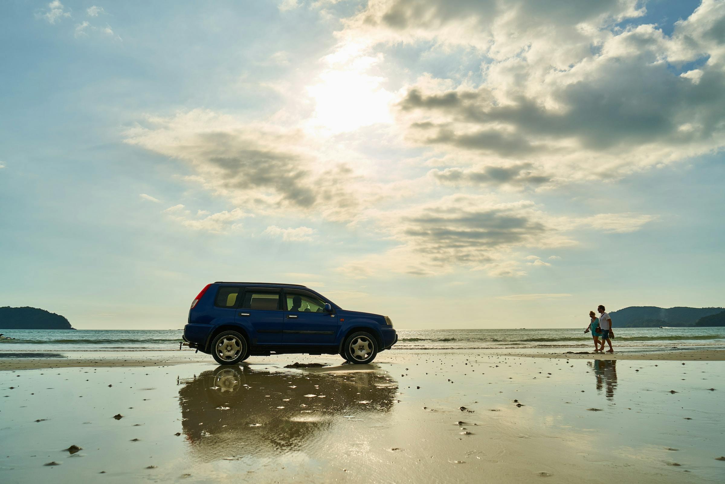 Two people walking from the right towards a blue SUV parked on a beach.