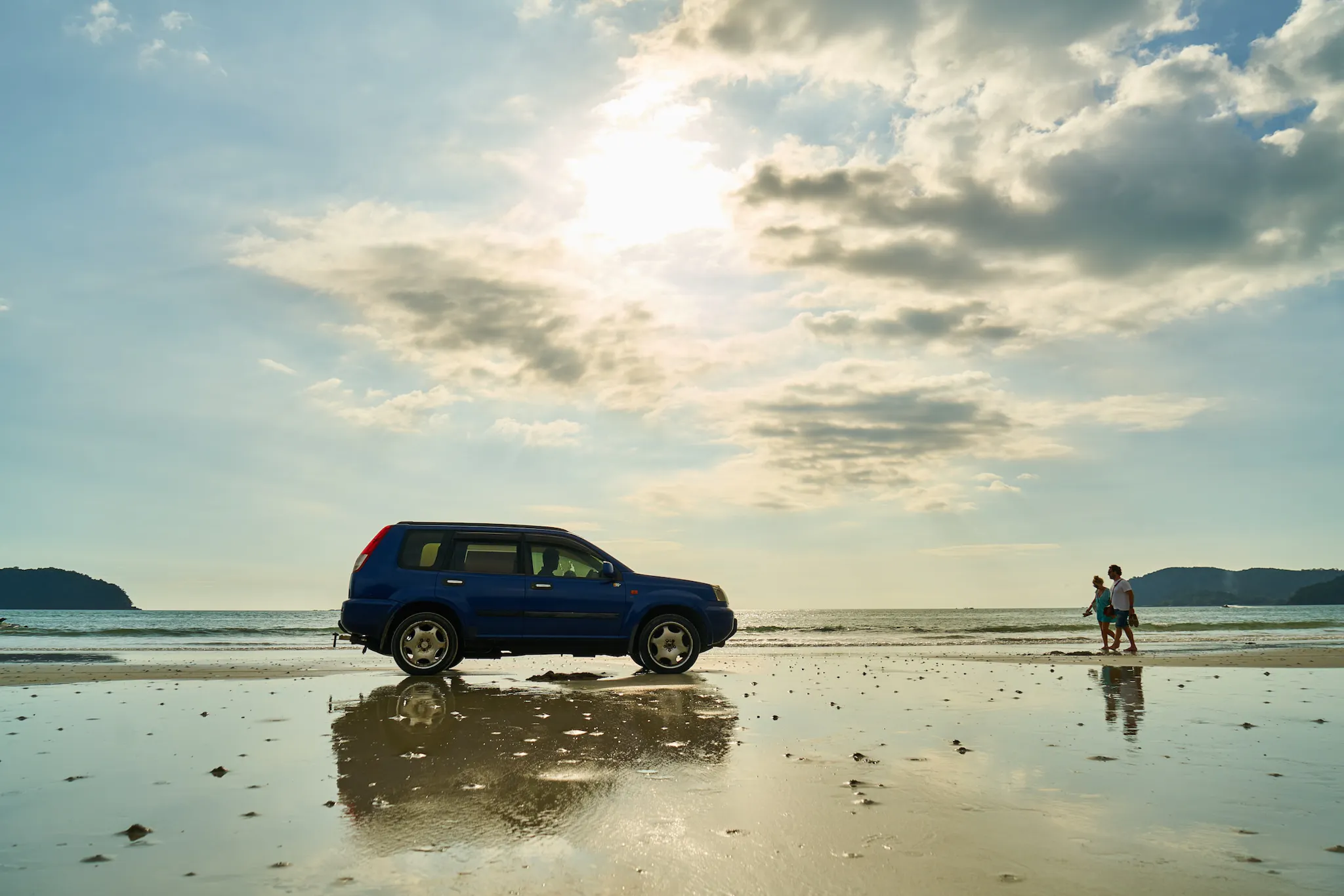 Two people walking from the right towards a blue SUV parked on a beach.