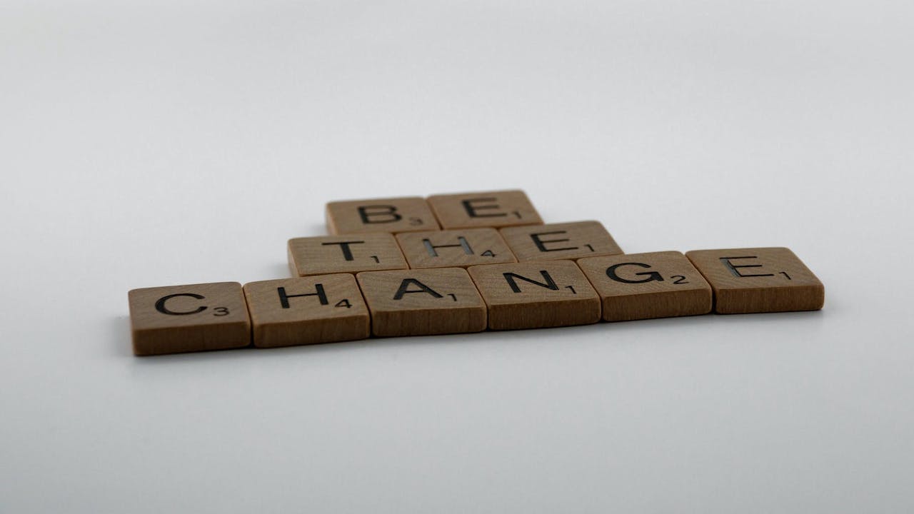 Wooden Scrabble pieces arranged to spell "Be the change" on a grey-and-white surface. 