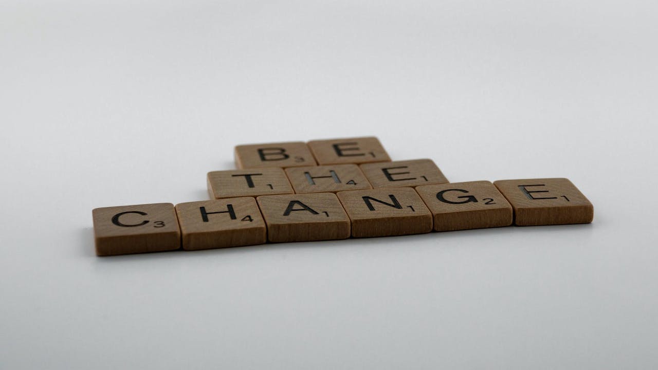 Wooden Scrabble pieces arranged to spell "Be the change" on a grey-and-white surface.