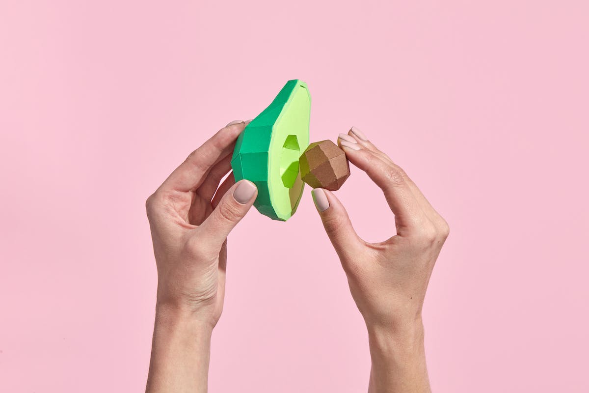 The hands of the woman hold a half of a ripe avocado and an avocado pit made with paper.