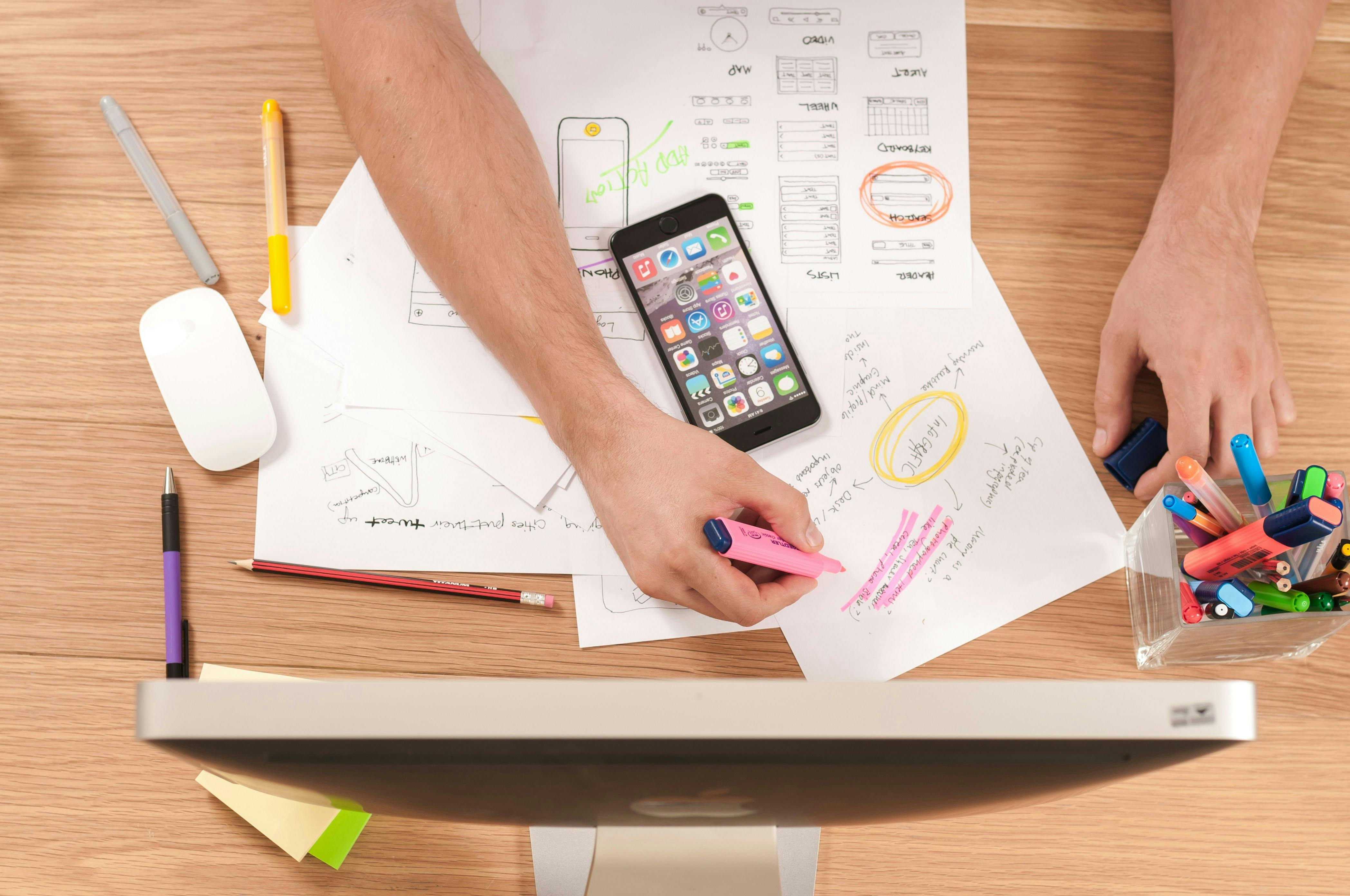 A picture shot from above, showing a computer screen, a mobile phone and a person's hands who is drawing something with a pink marker on a paper.