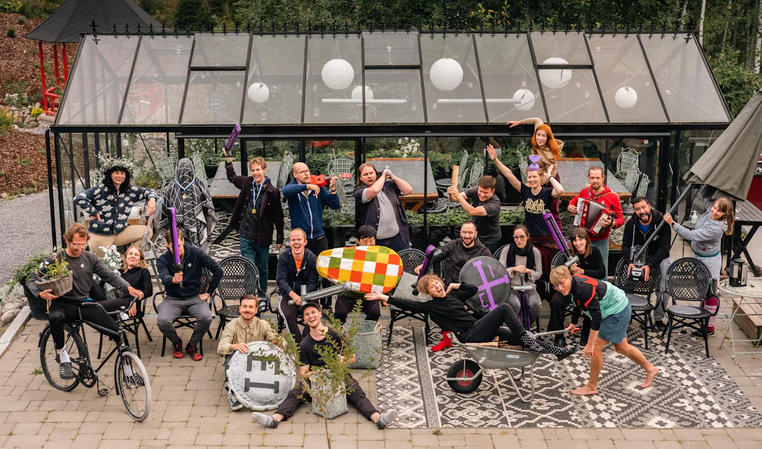 Photo of Sharetribe team in front of a greenhouse in funny poses and sporting various eclectic props, from plastic shields and soft buffering swords to a wheelbarrow, a harmonica, and a blender. 