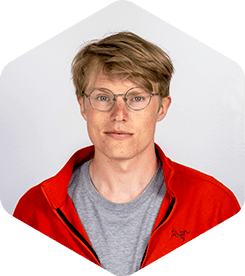 Tom is has a slight smile on his face. He's wearing classic round glasses and a sporty, red, zip-up sweatshirt over a grey t-shirt.