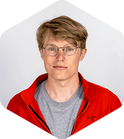 Tom is has a slight smile on his face. He's wearing classic round glasses and a sporty, red, zip-up sweatshirt over a grey t-shirt.