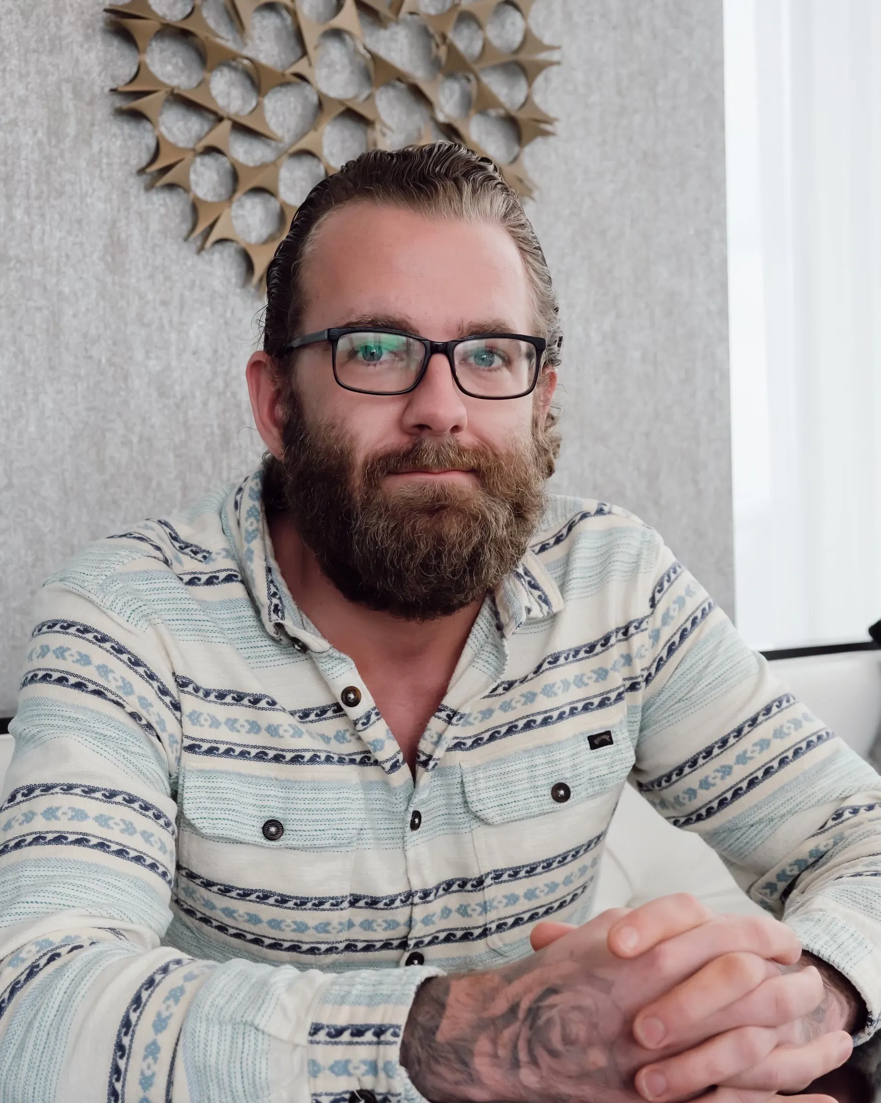 A bearded man with glasses, wearing a patterned long-sleeve shirt, sits at a table with his hands folded in front of him.