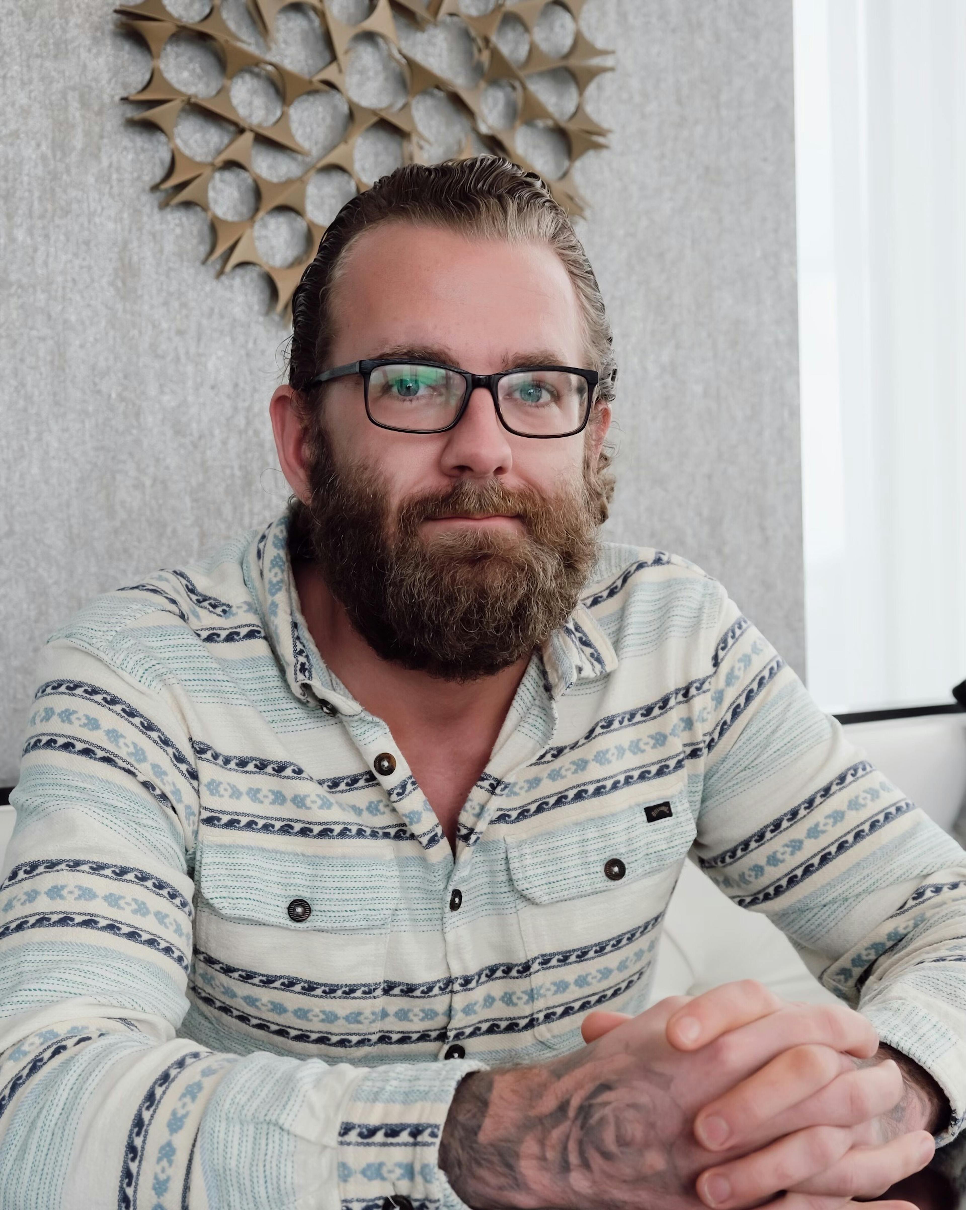 A bearded man with glasses, wearing a patterned long-sleeve shirt, sits at a table with his hands folded in front of him. 