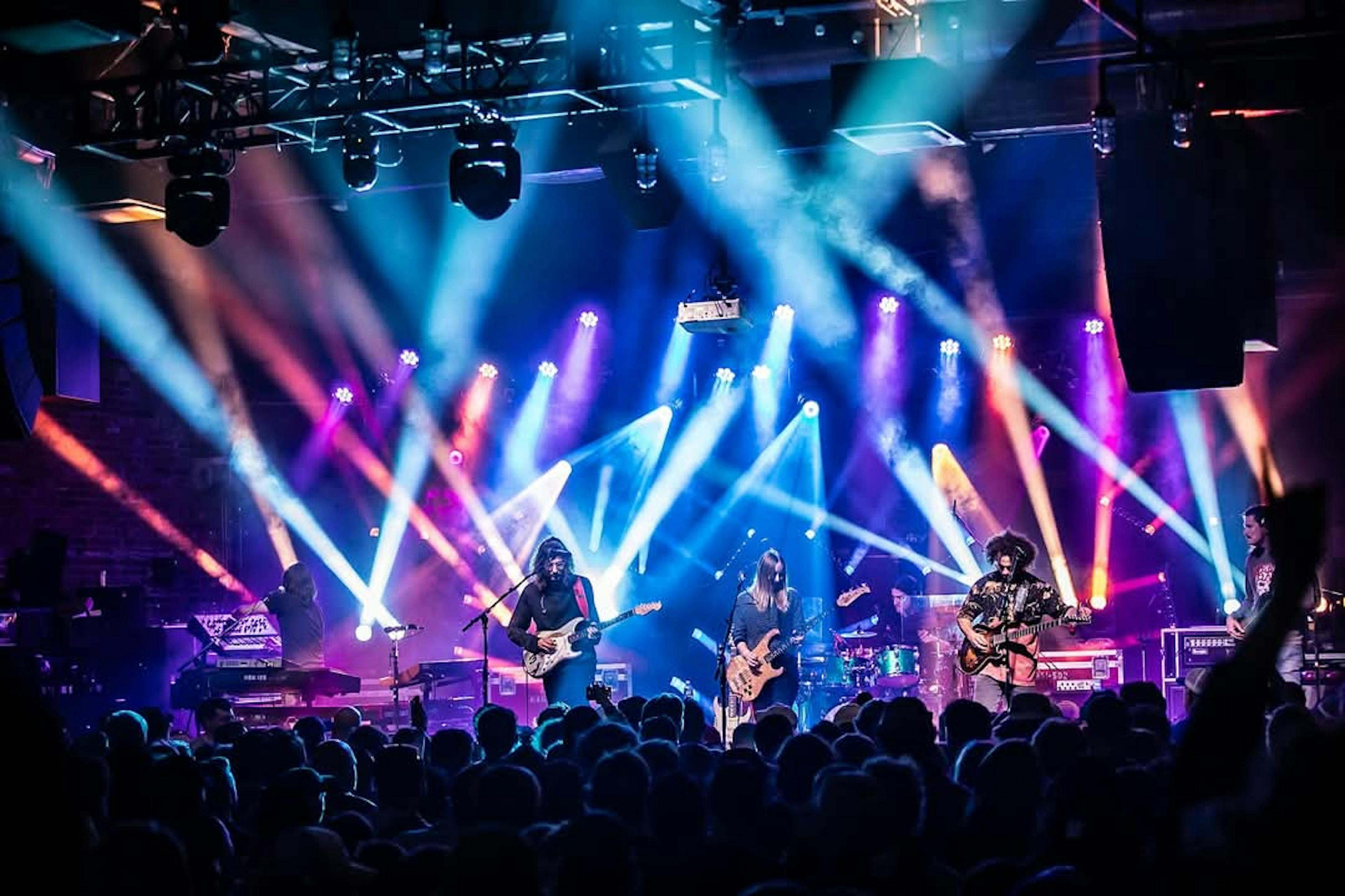A band playing at a concert stage with lots of lights in blue, orange, and purple hues and some rigging visible at the top of the photo.