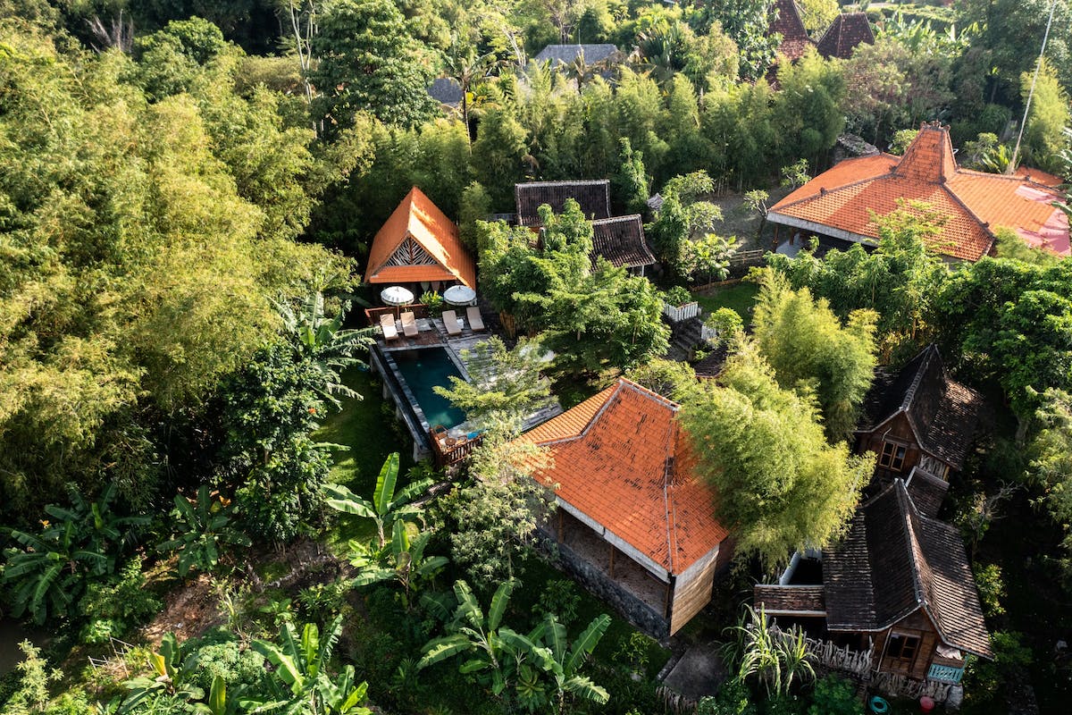 Birds-eye view of a holiday resort surrounded by lavish greenery: buildings with roofs of orange tile, bungalow-style huts, and a pool with sunchairs and parasols.