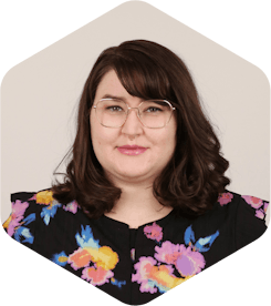 Amanda has dark, medium-lengthed brown hair, glasses, and a black floral shirt. She is smiling at the camera.