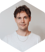 Picture of Aleksi, who has short, curly brown hair is looking directly at the camera with a slight smile. They are wearing a light grey t-shirt, a thin silver chain necklace, and a small hoop earring in their left ear. The background is plain and neutral, and the photo is cropped in a hexagonal shape.