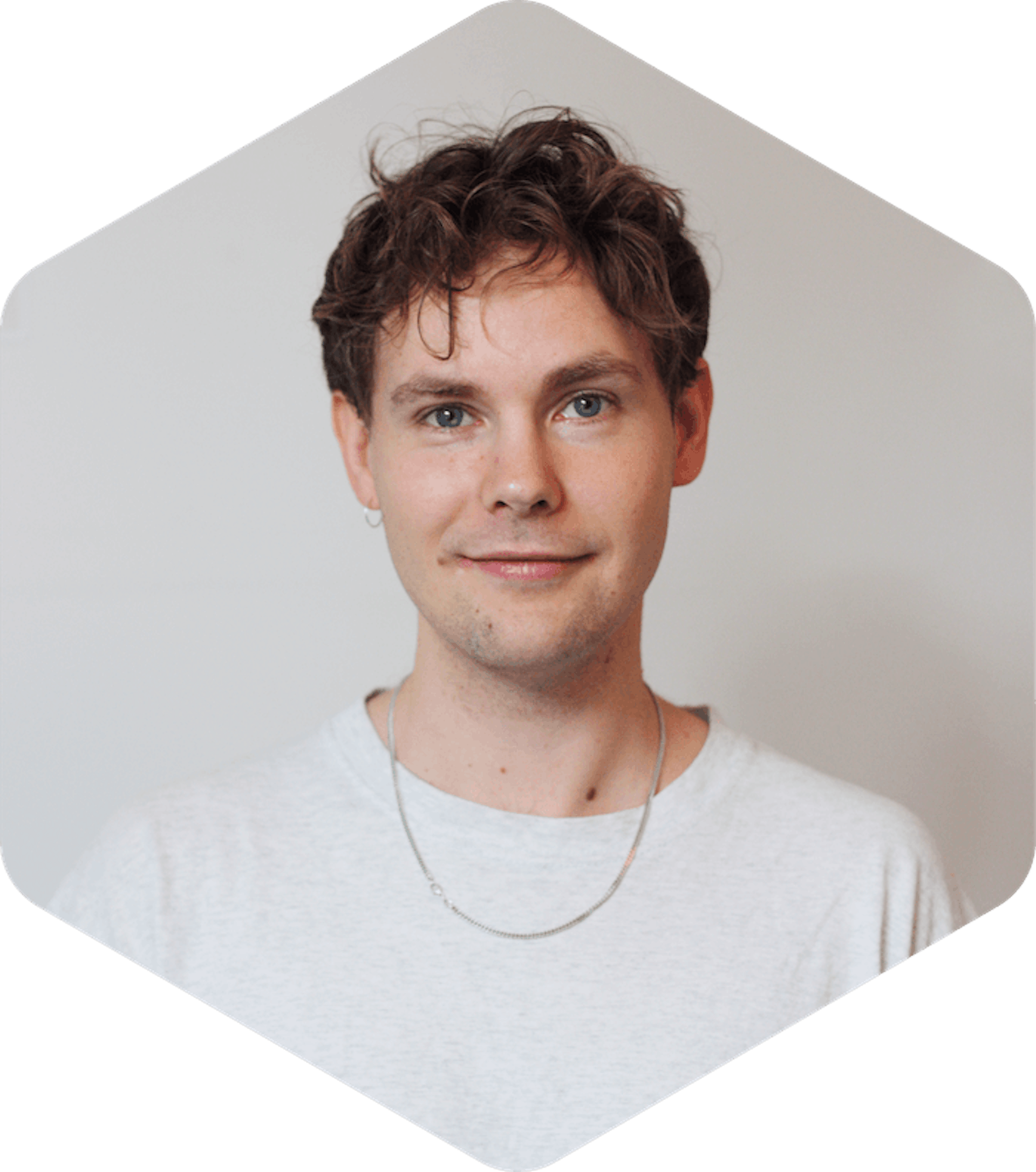 Picture of Aleksi, who has short, curly brown hair is looking directly at the camera with a slight smile. They are wearing a light grey t-shirt, a thin silver chain necklace, and a small hoop earring in their left ear. The background is plain and neutral, and the photo is cropped in a hexagonal shape.