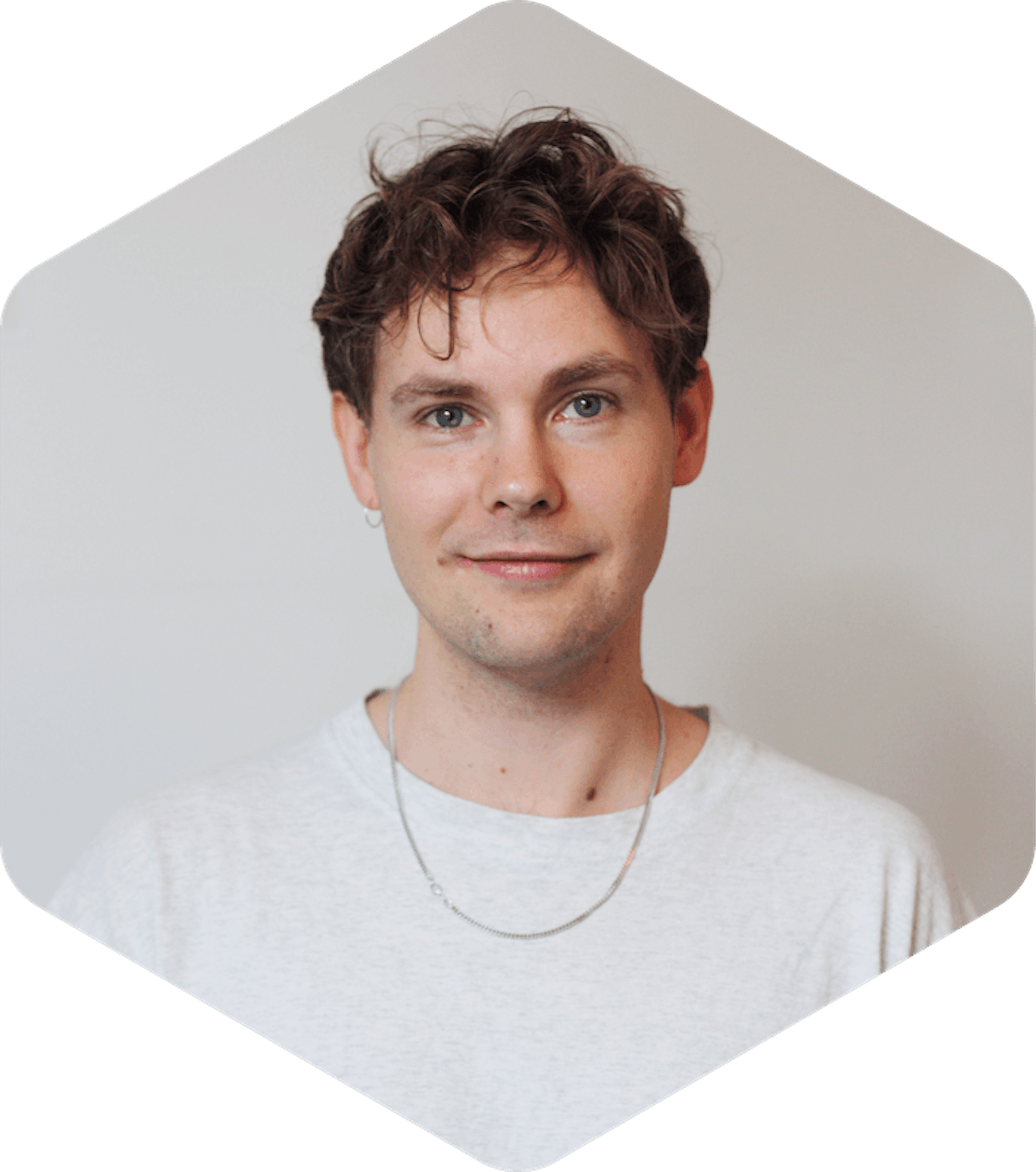 Picture of Aleksi, who has short, curly brown hair is looking directly at the camera with a slight smile. They are wearing a light grey t-shirt, a thin silver chain necklace, and a small hoop earring in their left ear. The background is plain and neutral, and the photo is cropped in a hexagonal shape.