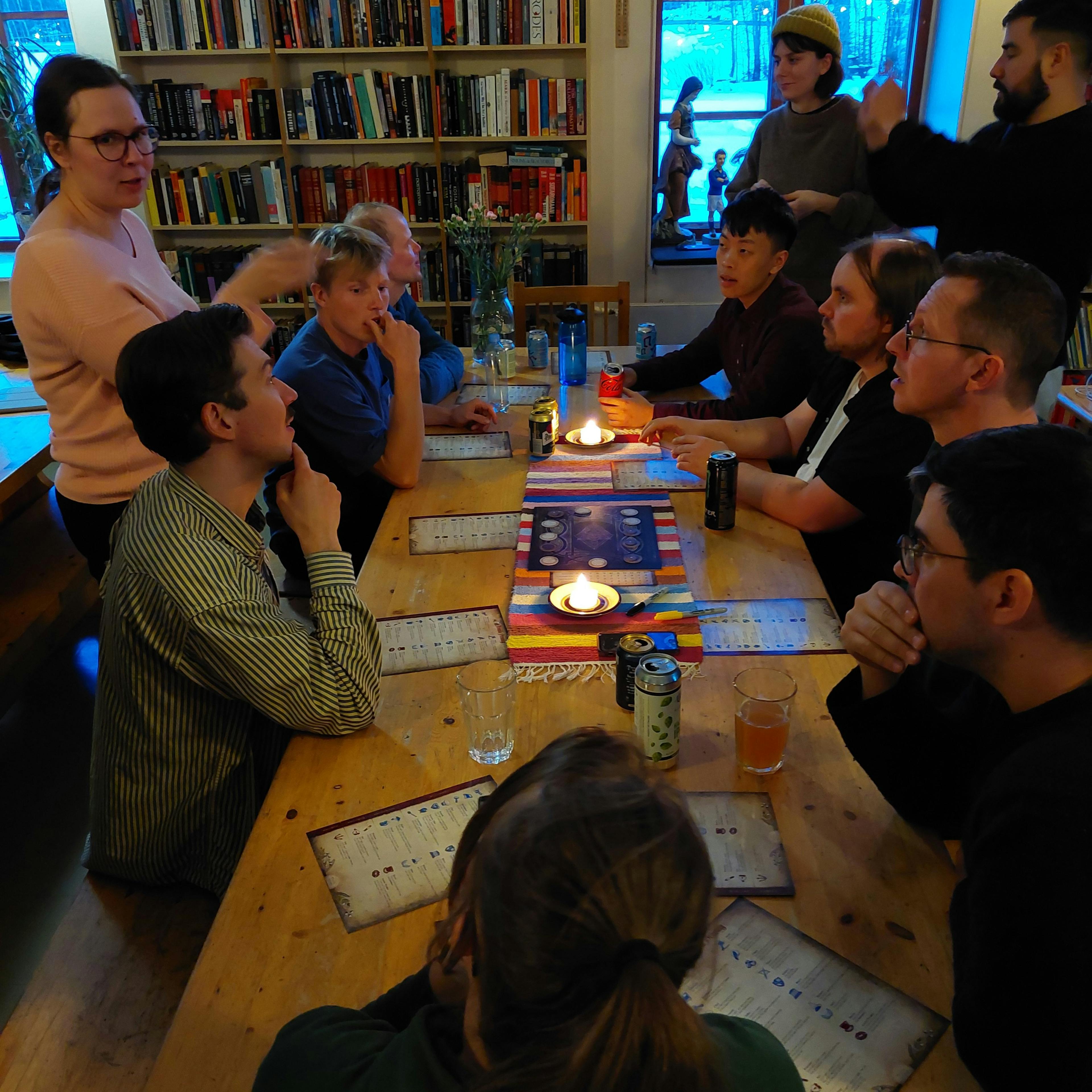 A group of people sit around a wooden table indoors, playing a board game by candlelight.