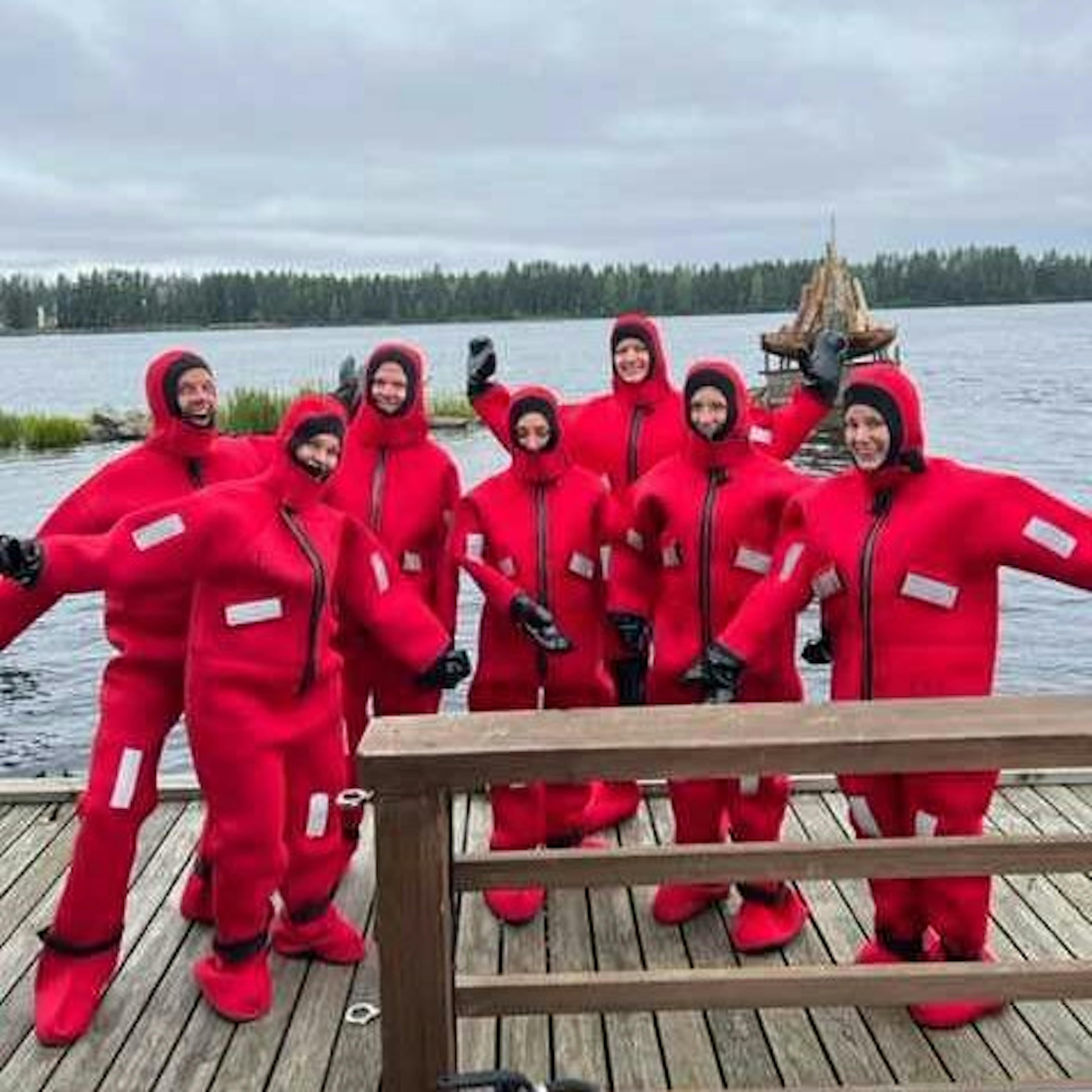 A group of people wearing bright red insulated suits pose together on a wooden dock by a lake, smiling and waving.