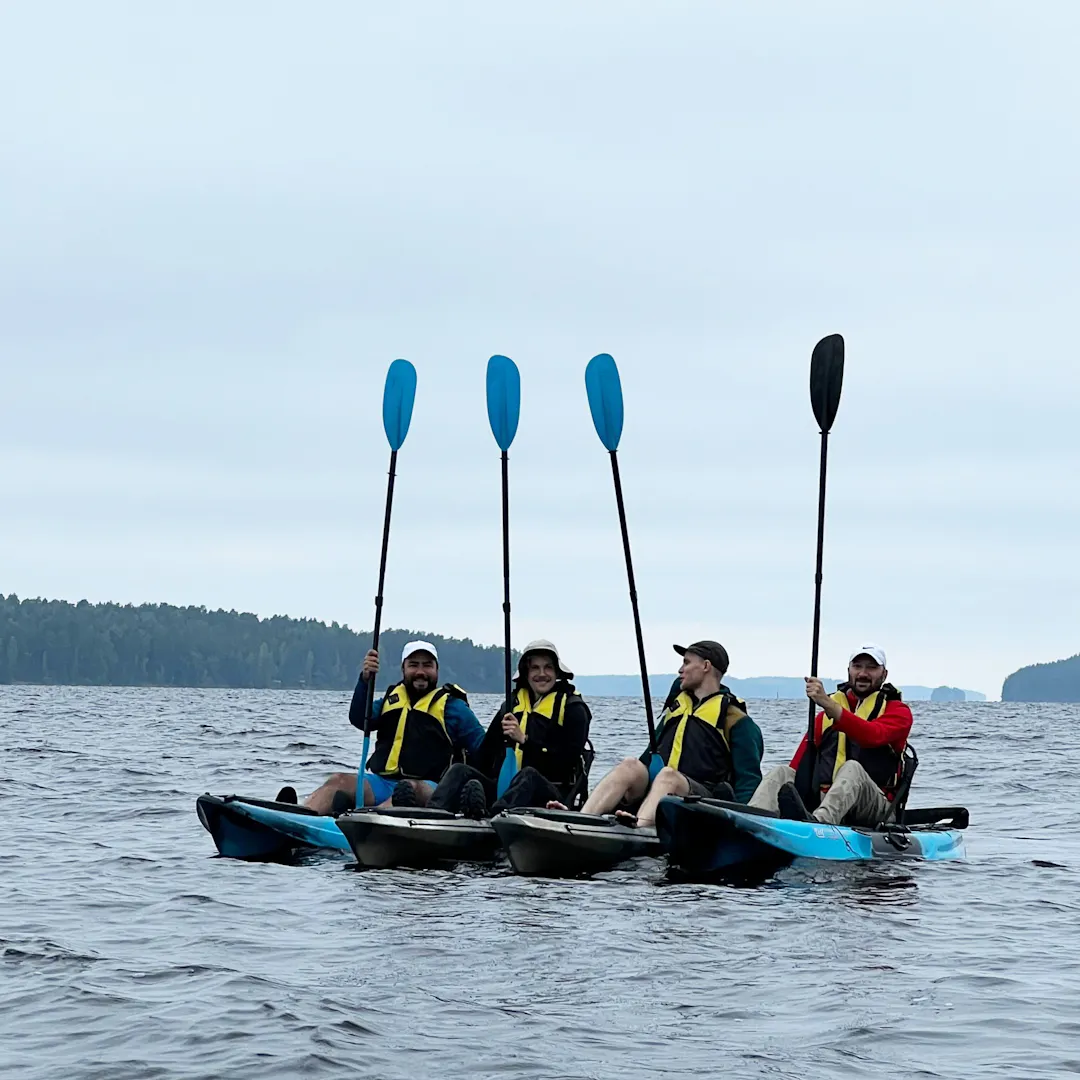 Four people in life vests sit in individual kayaks on open water, holding paddles upright and smiling toward the camera.