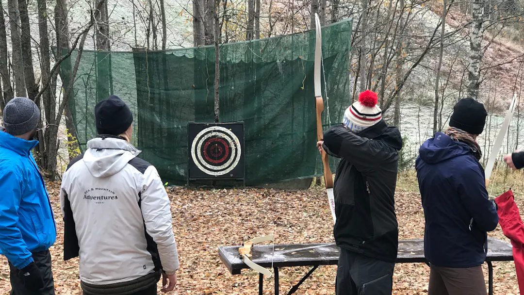 A group of people practicing archery outdoors in a forest, aiming at a target with bows.
